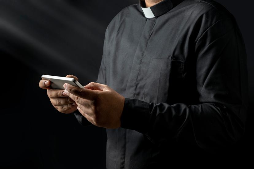Young priest holding handphone, texting something isolated on black background.